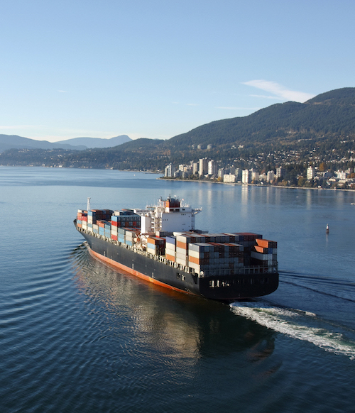 A large cargo ship sets off to sea with the shoreline dotted with buildings shown in the distance. This image is representative of the maritime injury services offered by MMRBH Law Office.