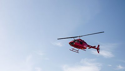 A red helicopter flies high in the sky. This photo serves as a header image for the article "What to Do After Your Loved One is in a Helicopter Crash" by MMRBH law office.