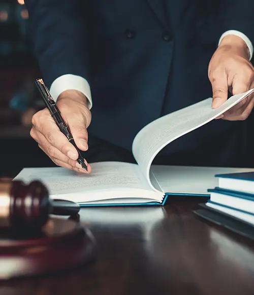 A vertical photo from the chest down of a person in a suit writing in a notebook. There is a gavel and sounding block, scale of justice and various books also sitting on the table. This photo is meant to represent MMRBH Law Office's mass tort lawsuit services.